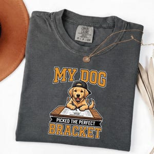 Può includere: T-shirt Comfort Colors grigio scuro con la scritta "MY DOG PICKED THE PERFECT BRACKET" in arancione e bianco. La grafica presenta un golden retriever con un berretto da baseball, seduto a una scrivania e con una matita in mano.