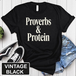 May include: A black short-sleeved t-shirt featuring the white text "Proverbs & Protein" printed in the centre of the chest. The garment has a crew neckline and is shown alongside a pair of blue denim shorts and black and white low-top trainers.