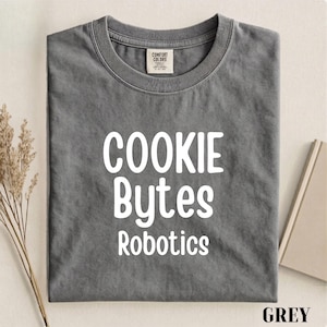 May include: A charcoal grey short-sleeve t-shirt featuring the white text COOKIE BYTES Robotics printed on the front. The shirt has a crew neckline and a Comfort Colors brand label visible on the inside of the collar area.