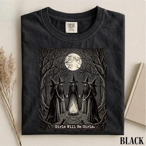 May include: A black t-shirt featuring a graphic print of five figures in pointed hats standing in a circle around a campfire in a dark forest under a full moon. The text "Girls Will Be Girls." is printed at the bottom of the graphic design.