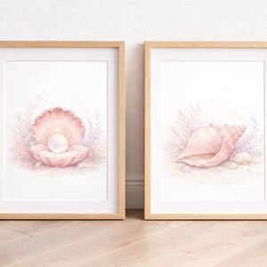 Könnte beinhalten: Zwei gerahmte Aquarelldrucke. Einer zeigt eine rosa Muschel mit einer Perle, der andere eine Muschel. Beide sind vor einem weichen, neutralen Hintergrund mit hellrosa und violetten Akzenten. Die Rahmen sind aus hellem Holz.