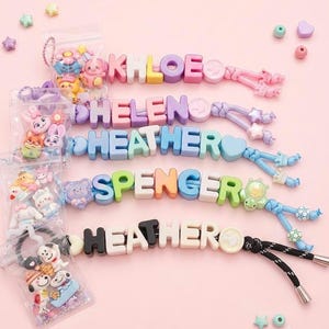 May include: Five personalized keychains featuring colorful block letters spelling KHLOE, HELEN, HEATHER, SPENCER, and HEATHER. Each keychain includes decorative character beads, elastic cords, and small plastic bags containing additional assorted charms.