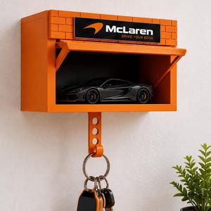 Op de afbeelding: Een oranje wanddisplay met een bakstenen patroon aan de bovenkant en een McLaren-logo met de tekst "McLaren DRIVE YOUR EDGE". In het open compartiment staat een donkergrijze modelauto. Een sleutelhanger met een McLaren-fob hangt aan de onderkant.