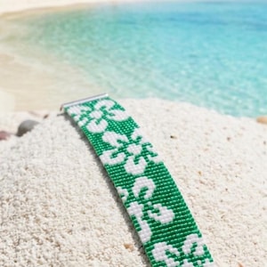 Könnte beinhalten: Ein Perlenarmband mit einem sich wiederholenden weißen Blumenmuster auf leuchtend grünem Hintergrund. Das Armband ist mit silberfarbenen Metallendstücken und einer passenden silberfarbenen Verlängerungskette mit rundem Verschluss versehen.