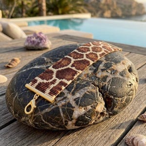 Könnte beinhalten: Ein Armband mit Perlenmuster im Giraffenmuster in Braun- und Cremetönen, das auf einem dunklen, strukturierten Stein liegt. Das Armband hat goldfarbene Metallenden und einen Verschluss. Der Hintergrund umfasst einen Pool und ein Holzdeck.