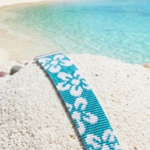 Könnte beinhalten: Ein breites Perlenarmband mit einem sich wiederholenden weißen Blumenmuster auf leuchtend türkisfarbenem Hintergrund. Das Armband ist mit silberfarbenen Metallendstücken und einer passenden silberfarbenen Verlängerungskette mit Ringverschluss versehen.