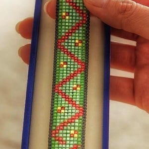 Könnte beinhalten: Ein rechteckiges Perlenarmband mit grünem Hintergrund, rotem Zickzackmuster und kleinen gelben Akzenten, das mit einem silberfarbenen Knebelverschluss versehen ist und in einer schmalen, aufrechten, blau ausgekleideten Schmuckschatulle liegt.