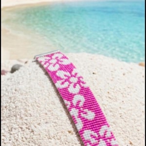 Könnte beinhalten: Ein breites Perlenarmband mit einem sich wiederholenden Blumenmuster aus leuchtend pinkfarbenen und strahlend weißen Glasperlen. Das Armband ist mit silberfarbenen Metallendstücken und einem feinen Kettengliedverschluss versehen, was ein strukturiertes Band ergibt.