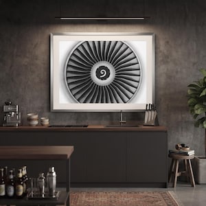 May include: A framed wall art print featuring a black and white close-up of a jet engine turbine with numerous radial blades and a central circular hub marked with a small spiral symbol, displayed in a modern kitchen setting above a dark countertop.