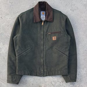 Puede incluir: Chaqueta de trabajo verde oscuro con cuello de pana marrón, cremallera y dos bolsillos delanteros. La chaqueta presenta un pequeño parche de cuero marrón y un pequeño bolsillo en el pecho. El logotipo de Carhartt es visible.