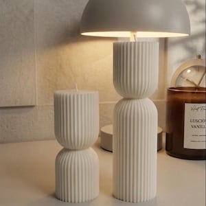 Peut inclure: Deux bougies piliers blanches et côtelées et une lampe blanche avec un abat-jour arrondi. Les bougies ont un design empilé. Une bougie marron avec du texte est visible en arrière-plan.