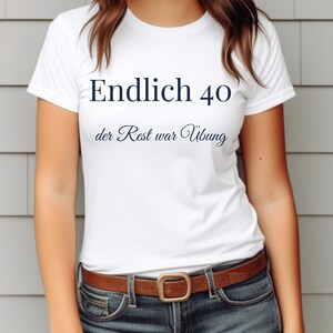 Könnte beinhalten: Weißes T-Shirt mit dem Text "Endlich 40" über "der Rest war Übung" in marineblauer Schrift. Das Shirt wird mit einer Jeans und einem braunen Ledergürtel getragen. Der Hintergrund ist eine graue Wand.