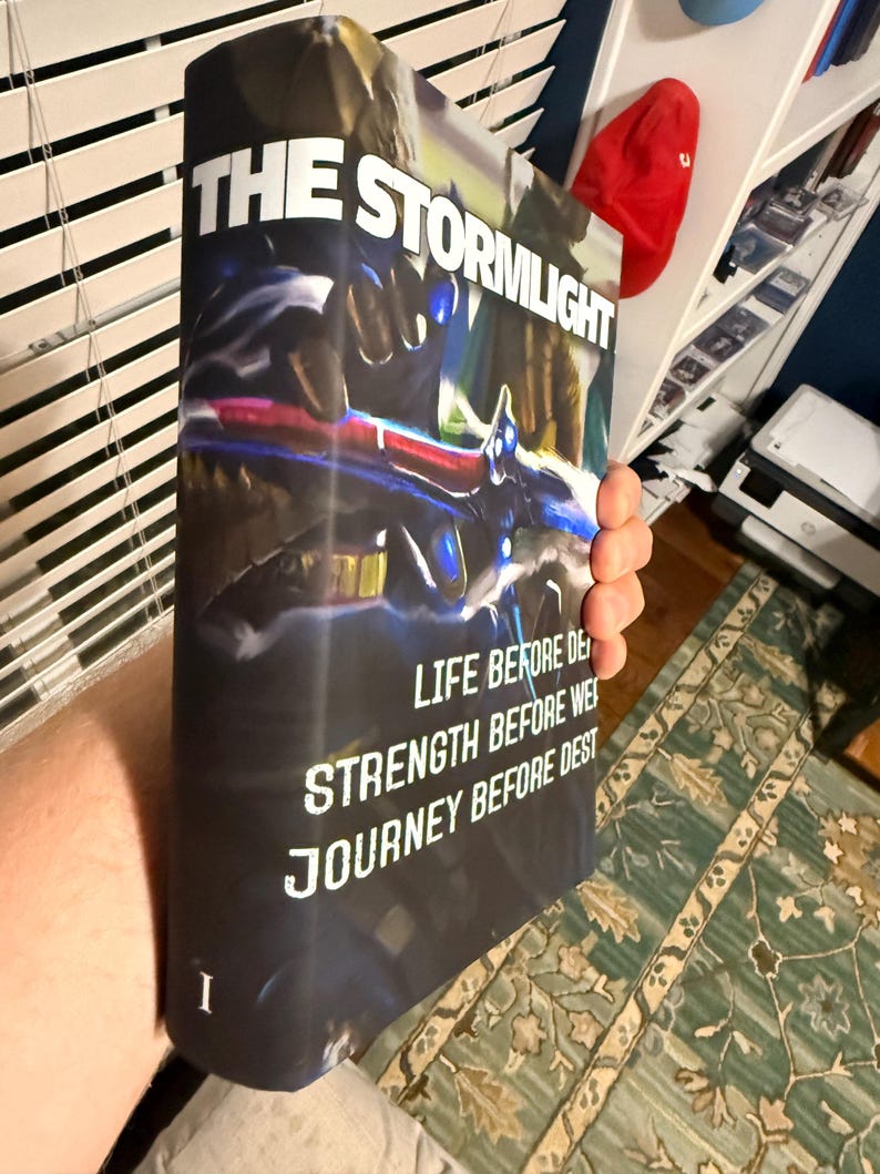 Puede incluir: Un libro de tapa dura titulado "The Stormlight Archive" con una cubierta oscura que presenta un dise&ntilde;o futurista en azul, rojo y blanco. El lomo del libro muestra el t&iacute;tulo y las frases "Life Before Death", "Strength Before Weakness" y "Journey Before Destination".