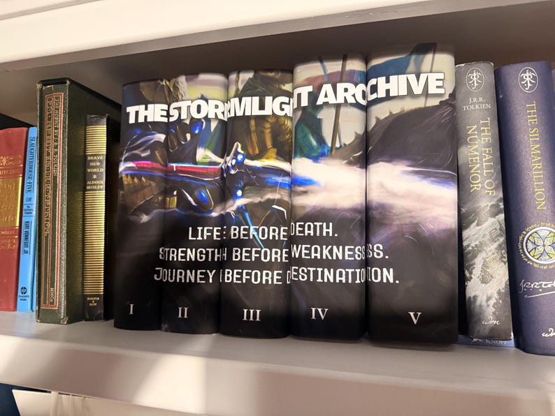 Puede incluir: Una estanter&iacute;a exhibe una colecci&oacute;n de libros de tapa dura. El enfoque principal est&aacute; en una serie titulada "THE STORMLIGHT ARCHIVE", con los vol&uacute;menes I a V visibles. Libros adicionales incluyen "The Fall of Numenor" y "The Silmarillion."
