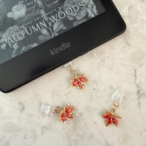 May include: Three gold-toned maple leaf charms with orange crystal accents, each attached to a clear plastic clip. The charms are positioned near a black Kindle e-reader displaying the book cover for Autumn Woods by Sorrowsong University.