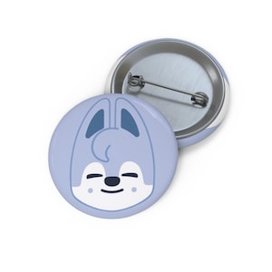 May include: Two round, light blue pinback buttons. One button features a cartoon bunny face with a smiling expression. The other button is face up, showing the pin back. The bunny face has dark blue ears and a black nose and mouth.