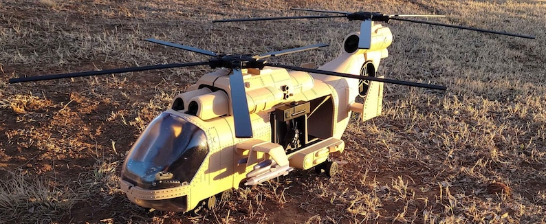 May include: A tan-colored military helicopter model featuring a clear cockpit canopy, black rotor blades, and an open side cargo door. The fuselage displays a light brown finish with visible mechanical details, including side-mounted missiles and a tail rotor.