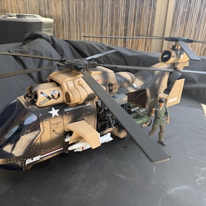 May include: A tan and black G.I. Joe toy helicopter featuring a dark cockpit, rotating rotor blades, and side-mounted missiles. The fuselage displays a white star emblem and the text G.I. JOE. A small action figure stands beside the open side door.