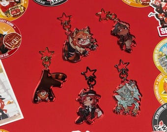 May include: Five acrylic keychains with star-shaped clasps featuring various illustrated characters, including a red panda, a person with glasses, a person in a hat, and a creature, arranged on a red surface alongside several circular stickers.