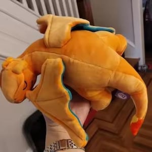 May include: Orange plush toy dragon with yellow wings and blue accents. The dragon has a long tail with a red tip. The toy is soft and cuddly, perfect for fans of fantasy creatures.