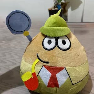 May include: A plush toy character in the shape of a tan potato with large black eyes, wearing a green hat and a red and yellow pipe. It holds a blue magnifying glass. The character has a white shirt and a red tie.