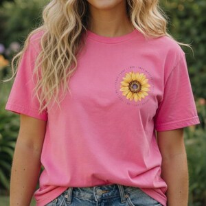 以下が含まれることがあります： 前面にヒマワリのグラフィックがプリントされたピンク色のTシャツ。ヒマワリは黄色で、中心は茶色で、周りにテキストがあります。このシャツはデニムショーツと組み合わせています。