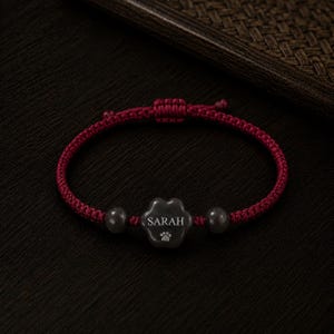 Könnte beinhalten: Burgunderfarbenes geflochtenes Armband mit einem Pfotenabdruck-Anhänger. Der dunkelgraue Anhänger trägt den Namen "SARAH" in weißer Schrift. Zwei graue Perlen flankieren den Anhänger. Das Armband ist am Verschluss geknotet.