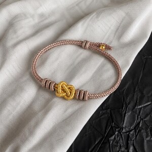 Könnte beinhalten: Ein Armband mit goldfarbenem Knotenanhänger und rosa Kordel. Das Armband hat ein zentrales Knotendesign und ist mit goldfarbenen Perlen verziert. Die Kordel ist hellrosa.
