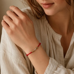 Könnte beinhalten: Ein rotes Armband aus Kordel mit einer zentralen silberfarbenen zylindrischen Perle, die auf jeder Seite von zwei kleineren goldfarbenen Metallperlen flankiert wird. Das Armband besteht aus einer dünnen, glatten roten Schnur.