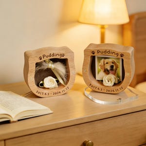 Könnte beinhalten: Zwei hölzerne Gedenkrahmen für Haustiere mit dem Namen "Pudding" und eingravierten Daten. Ein Rahmen zeigt ein Hundefoto mit einer weißen Rose, der andere enthält Fell und eine weiße Rose. Beide stehen auf einem Nachttisch aus Holz.