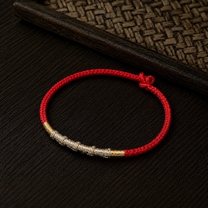 Könnte beinhalten: Ein rotes geflochtenes Armband mit goldenen und silbernen Akzenten. Das Armband hat eine Reihe silberfarbener Perlen und goldfarbener Bänder. Das Armband ist am Ende geknotet. Das Armband liegt auf einer schwarzen Oberfläche.