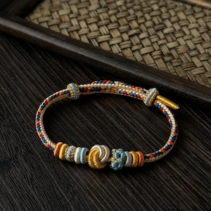 Può includere: Un braccialetto intrecciato con un motivo beige, rosso, blu e giallo. Il braccialetto presenta nodi e perline decorative nei toni dell'oro, blu e argento. Il braccialetto è esposto su una superficie di legno scuro.
