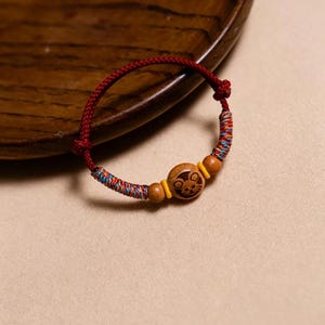 Op de afbeelding: Een rood gevlochten armband met een houten kraal met een gesneden dierenontwerp. De armband heeft kleurrijke geweven accenten en twee houten kralen aan weerszijden van de centrale kraal. De armband wordt gepresenteerd op een houten oppervlak.