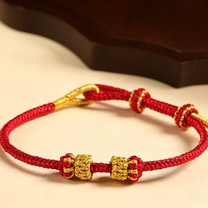 Könnte beinhalten: Rotes gewebtes Armband mit goldfarbenen Akzenten. Das Armband hat goldfarbene Perlen und einen Verschluss. Das Armband besteht aus einem roten gewebten Material und hat goldfarbene Metalldetails. Das Armband ist ein traditioneller Stil.