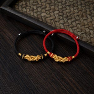 Könnte beinhalten: Zwei Armbänder: eines schwarz, eines rot. Jedes hat ein goldfarbenes Knotendesign in der Mitte. Die Armbänder haben verstellbare Kordeln und silberfarbene Endperlen. Der Hintergrund ist eine dunkle Holzoberfläche.