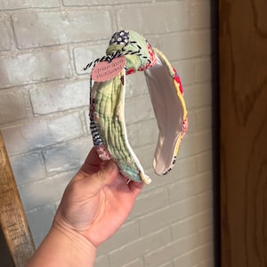 May include: A knotted headband crafted from multicolored quilted fabric featuring various patterns including green, red, and blue. A small pink tag attached with black and white twine displays the handwritten text green quilt headband.