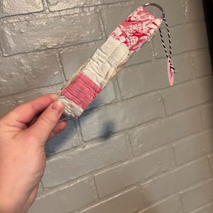 May include: A fabric keychain featuring a patchwork design with sections of white, pink, and a red and white patterned textile. The keychain includes a silver metal ring and a small black and white string with a circular pink tag attached to the end.