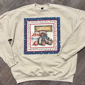 May include: A beige crewneck sweatshirt featuring a central square graphic panel. The graphic depicts a brown teddy bear with a blue ribbon, a basket of red apples, and a sign that reads Fresh Apples 5c, all framed by a blue floral border.