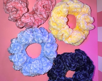 May include: Four plush scrunchies in shades of pink, yellow, light blue, and navy blue. The scrunchies are made of a soft, velvety material and are arranged against a pink background. These hair accessories are perfect for adding a pop of color.