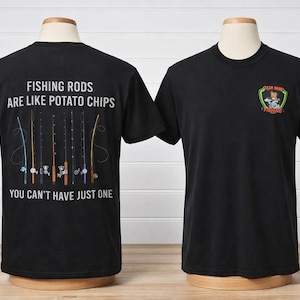 Puede incluir: Camisetas negras con gráficos de pesca. Una camiseta muestra el texto "FISHING RODS ARE LIKE POTATO CHIPS, YOU CAN'T HAVE JUST ONE" con ilustraciones de cañas de pescar. La otra tiene un logotipo que dice "TEAM HARD FISHING".
