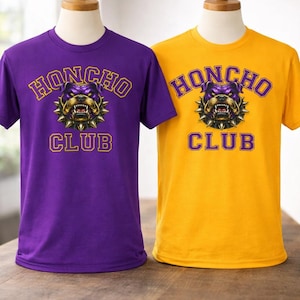 Op de afbeelding: Twee t-shirts, &eacute;&eacute;n paars en &eacute;&eacute;n goud, gepresenteerd op mannequins. Elk shirt heeft de tekst "HONCHO CLUB" boven een bulldog-afbeelding. De bulldog is paars en goud met een spiked kraag. De tekst is goud.