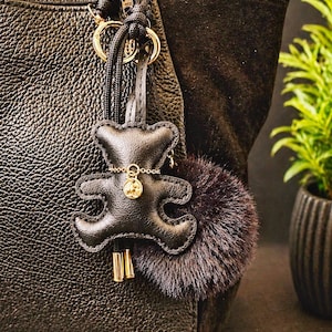 Könnte beinhalten: Schwarze Lederhandtasche mit einem Teddybär-Schlüsselanhänger und einem dunkelvioletten Fell-Pompon. Der Schlüsselanhänger hat eine goldene Kette und einen kleinen goldenen Anhänger. Die Tasche hat eine strukturierte Oberfläche und goldfarbene Hardware.