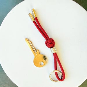 Könnte beinhalten: Ein goldener Schlüssel und ein roter Schlüsselanhänger mit Knoten und goldenem Ring. Der Schlüsselanhänger hat goldene Akzente oben. Die Artikel liegen auf einer weißen, kreisförmigen Oberfläche.