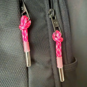 Könnte beinhalten: Ein Rucksack aus schwarzem, strukturiertem Stoff mit zwei silbernen Metall-Reißverschlussanhängern. Jeder Anhänger ist an einer leuchtend pinken, geknoteten Kordel mit einer silbernen, zylindrischen Metallspitze befestigt, die sich vom dunklen Stoff abhebt.