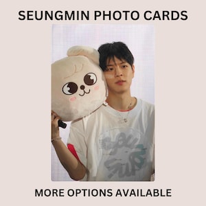 May include: A photo card featuring a person with dark hair wearing a white t-shirt, holding a light-colored plush toy with a cartoon face. The text SEUNGMIN PHOTO CARDS is at the top and MORE OPTIONS AVAILABLE is at the bottom of the image.