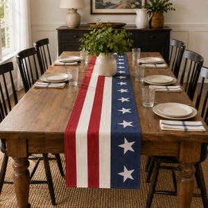 May include: A long rectangular table runner featuring a patriotic design with vertical red and white stripes on one side and a navy blue section with white stars on the other, laid across a wooden dining table with place settings.