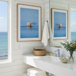 May include: A bathroom with a coastal theme. Two framed art prints with a blue background and seashells hang above a white countertop with a sink. A woven basket and a bowl of seashells sit on the counter. White towels are stacked on a shelf below.