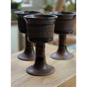 May include: Three ceramic goblets with a dark brown, glossy glaze and a wide, textured band around the upper bowl. Each vessel features a flared rim, a slender stem, and a circular base, showcasing a consistent earthy color palette throughout.