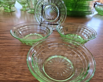 May include: A collection of vintage green glass bowls and plates. The bowls have a ribbed design around the sides and a decorative pattern in the center. The glassware is arranged on a wooden surface.