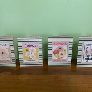 Peut inclure: Quatre cartes décoratives avec des illustrations florales et du texte. Les cartes présentent des images de renoncules, de cosmos, de zinnias et de thym. Chaque carte a une bordure rayée et des étiquettes de texte telles que "delicate beauty" et "fresh herbs".